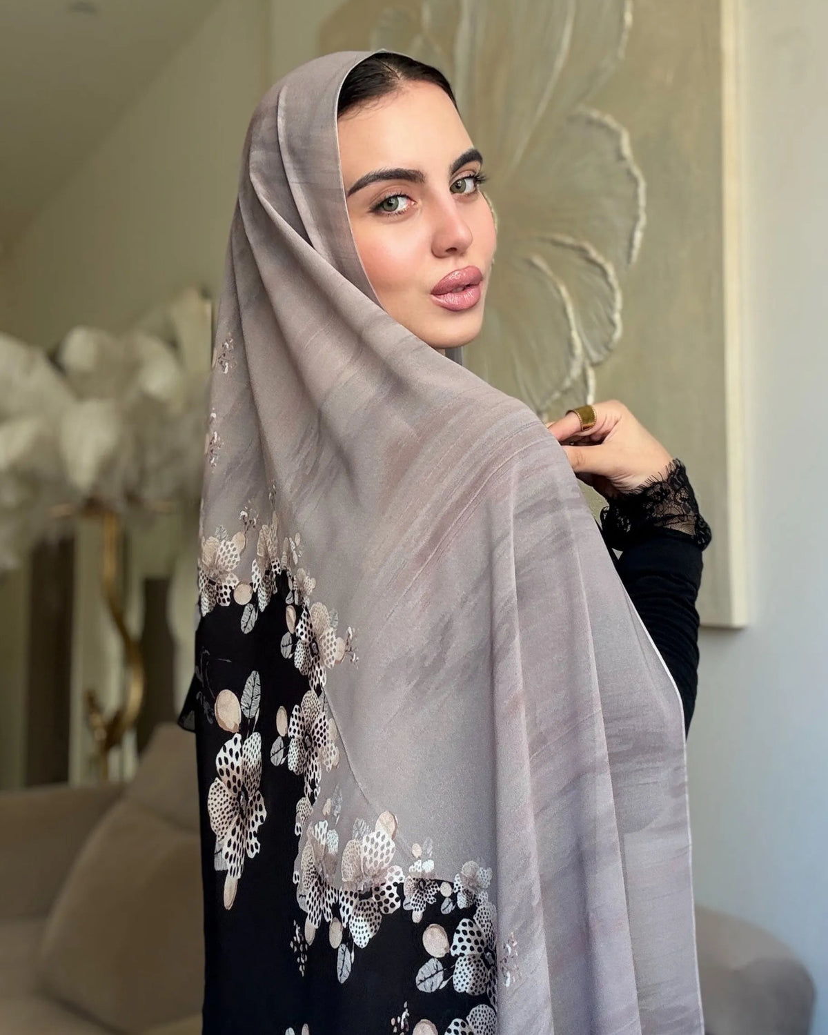 A woman wearing a light gray hijab with white floral embroidery and a black dress, standing in a modern living room setting.
